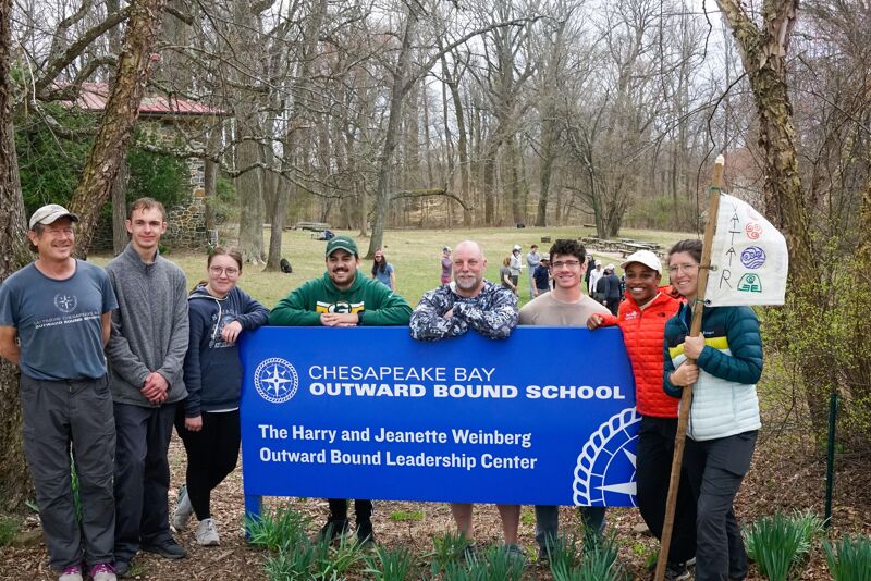 A group of nine people stand behind a blue sign that reads "Chesapeake Bay Outward Bound School, The Harry and Jeanette Weinberg Outward Bound Leadership Center". They are in a wooded area with a building visible in the background. The people are diverse in age and ethnicity, and they are all smiling. One person holds a flag with some drawings on it.